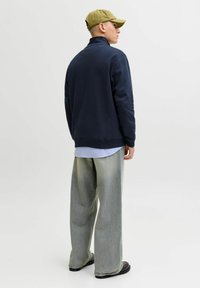 Tumed heeringi särk kõrge kaelaga, helesinine särk all, kombineeritud laia halli pükstega ja mustade kingadega; isikul on peas oliivroheline müts.