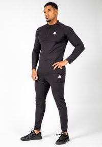 Zwart atletisch shirt met lange mouwen en ritssluiting aan de kraag, voorzien van een logo op de borst, gecombineerd met bijpassende strakke broek en zwarte sportschoenen.