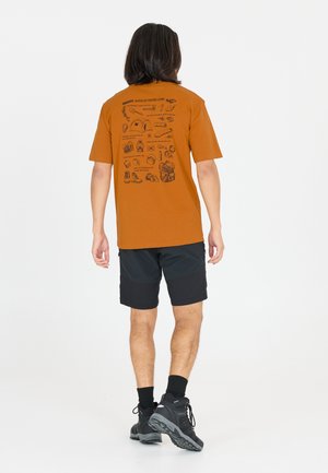 Persoon draagt een oranje t-shirt met zwarte illustraties en tekst gerelateerd aan wandelen op de achterkant, zwarte korte broek, zwarte sokken en zwarte wandelschoenen.