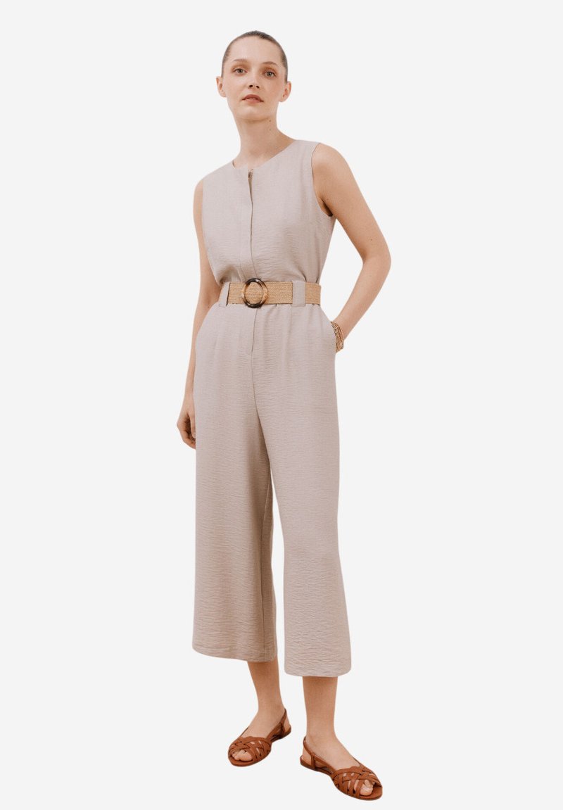 Beige jumpsuit med ärmlös design, texturerad tyg, knappstängning, vida avslutade ben och vävd bälte med rund spänne. Bruna sandaler.