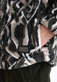 Fleece jack met een abstract zwart, bruin en crème patroon; bevat een zijzak en logo-tag. Getextureerd, zacht materiaal.
