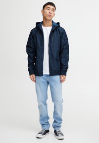 Laivasilakanvärinen kevyt takki hupparilla, vetoketjullinen etuosa ja elastiset hihansuut, yhdistettynä vaaleansinisiin farkkuihin sekä musta-valkoisiin lenkkareihin.