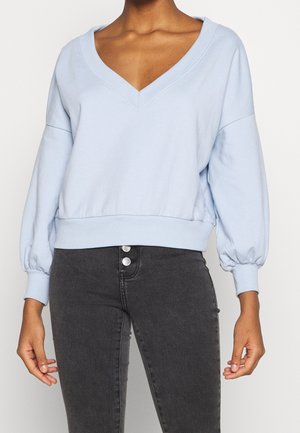 Persoon draagt een lichtblauw cropped sweatshirt met V-hals en lange mouwen en een hooggetailleerde donkergrijze spijkerbroek met twee zichtbare knopen.