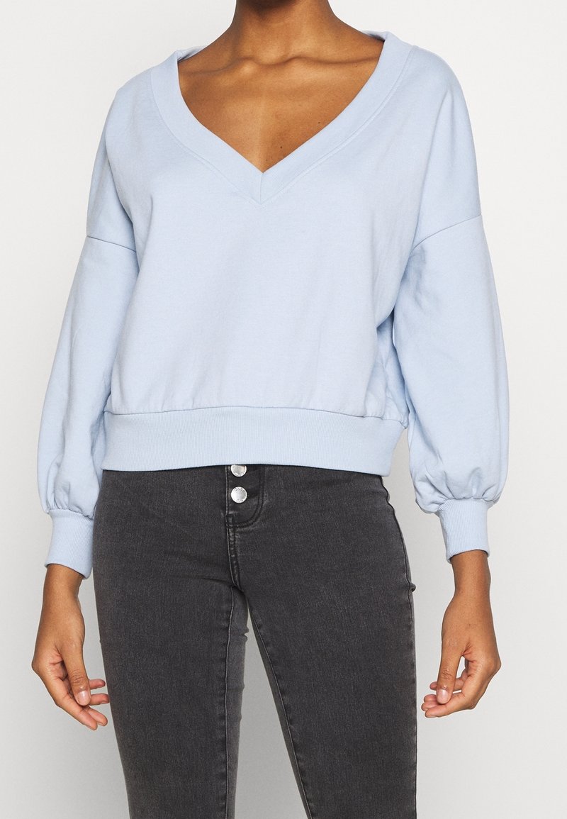 Personne portant un sweat-shirt court bleu clair à col en V et manches longues, ainsi qu’un jean taille haute gris foncé avec deux boutons visibles.