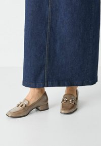 Bruna lackläders loafers med låg klack och dekorativt guldhardware. Bärs med en lång mörk denimkjol.