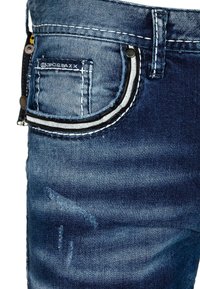 Jean en denim avec un délavage bleu foncé, présentant des coutures contrastées, un contour de poche décoratif et une texture légèrement distroyée.