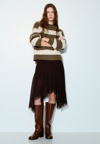 Pull en tricot à rayures crème et olive, superposé sur une jupe asymétrique marron à bordure en dentelle, complété par de longues bottes en cuir marron.