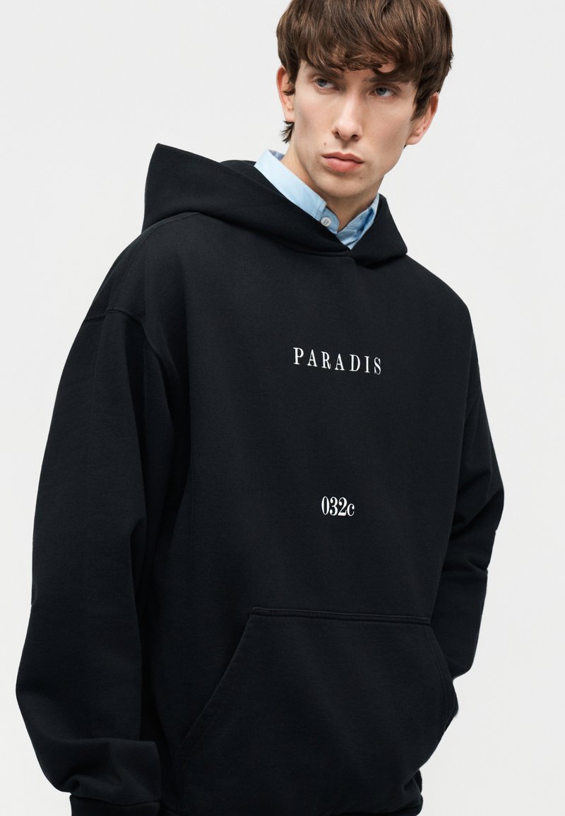Jeune homme portant un sweat à capuche noir avec les textes "PARADIS" et "032c", superposé sur une chemise à col bleu clair, regardant sur le côté.