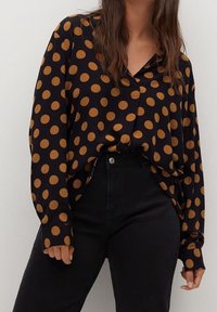 Mustapohjainen pitkähihainen paita, jossa on suuria ruskeita pilkkuja, V-mallinen kaula-aukko ja rento leikkaus. Yhdistetty korkeavyötaisien mustien housujen kanssa.