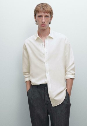 Light beige button-up shirt with rolled sleeves, paired with dark gray pinstriped trousers, worn with hands in pockets against a neutral background.