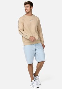 Sudadera beige con texto negro en las mangas, combinada con pantalones cortos de mezclilla azul claro. La modelo lleva zapatillas atléticas blancas y está posando de pie.