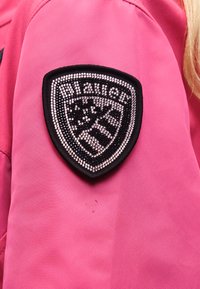 Patch en forme de bouclier orné de strass noirs et blancs avec le texte « Blauer » sur le bras supérieur d'une veste rose vif.