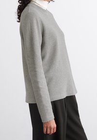 Maglione grigio chiaro con texture, maniche lunghe e collo a giro, indossato sopra un dolcevita bianco, abbinato a pantaloni scuri.