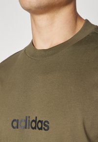 Olivově zelené bavlněné tričko s kulatým výstřihem a krátkými rukávy, na hrudi s černým logem "adidas". Hladká textury.
