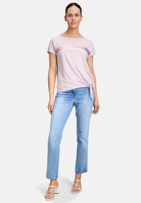 Jasnoróżowa koszulka z cekinowym poziomym paskiem, zestawiona z jasnoniebieskimi dżinsami. Koszulka ma związany bok i krótkie rękawy.