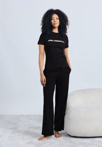 Tricou negru din bumbac cu logo-ul „KARL LAGERFELD”, asortat cu pantaloni negri. Modelul stă pe un covor bej lângă o canapea albă cu textură.