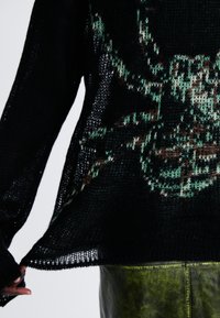 Zwarte gebreide trui met een losse weving en een groen en bruin spinnenontwerp. Zichtbare zoom en lange mouwen. Draag over een groene leren rok.