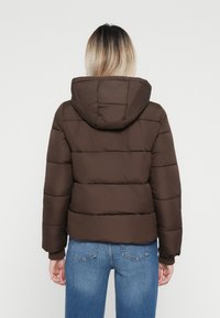 Veste rembourrée marron avec capuche, dotée de panneaux matelassés et de poignets côtelés, associée à un jean en denim bleu clair.