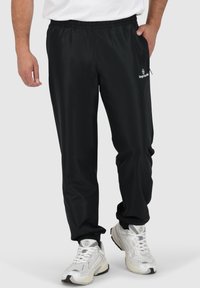 Schwarze Sporthosen mit einem elastischen Bund, seitlichen Taschen und slim fit. Mit einem Logo auf dem linkenOberschenkel. Glatter, leichter Stoff.