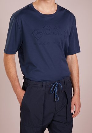Mężczyzna noszący granatowy t-shirt Hugo Boss z wytłoczonym logo i pasujące granatowe spodnie ze niebieskim sznurkiem, ręce w kieszeniach.