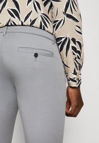 Light grey trousers with a smooth texture, featuring a back pocket with a black button. Paired with a patterned leaf print blouse.