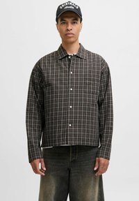 Een model in een geruite overhemd met knopen en donkere jeans staat tegen een eentonige achtergrond, kijkt recht in de camera.