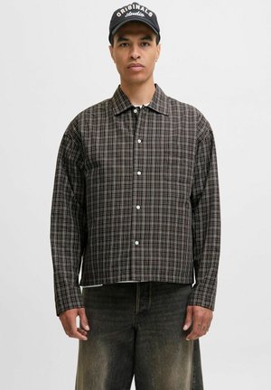 A model wearing a checkered button-up shirt and dark jeans stands against a plain background, looking directly at the camera.
