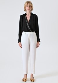 Schwarze Bluse mit silbernem Reißverschlussdetail, kombiniert mit maßgeschneiderten weißen Hosen und flachen Schuhen mit goldenen Akzenten, präsentiert eine schlanke, moderne Silhouette.