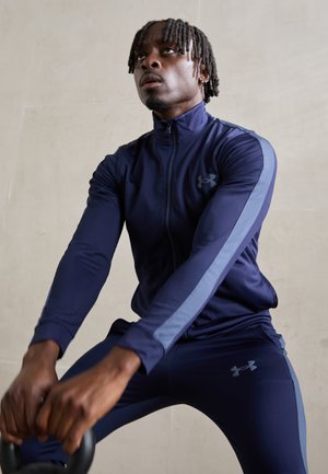 Tuta sportiva blu navy con strisce laterali azzurre, realizzata in tessuto liscio. Inquadratura ravvicinata di mani che afferrano un kettlebell, espressione concentrata.