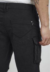 Black cargo shorts made of denim, featuring a side pocket, back pockets with a chevron design, and a smooth texture.