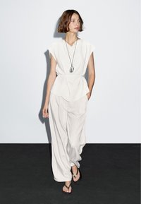 Hvid, kortærmet top med peplum-design, parret med løse, lyse bukser og flip-flops; halskæde med en stenpendant.