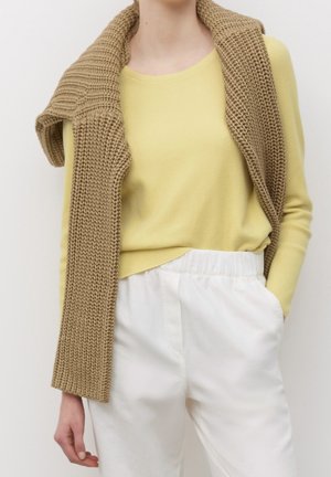 Person wearing yellow long-sleeve top and white pants, with tan knitted sweater draped over shoulders, hand in pocket.