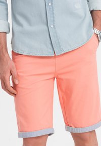 Camicia azzurra chiara da uomo e pantaloni corti arancione corallo con risvolti, caratterizzati da una texture liscia e un design casual. Orologio argento al polso.