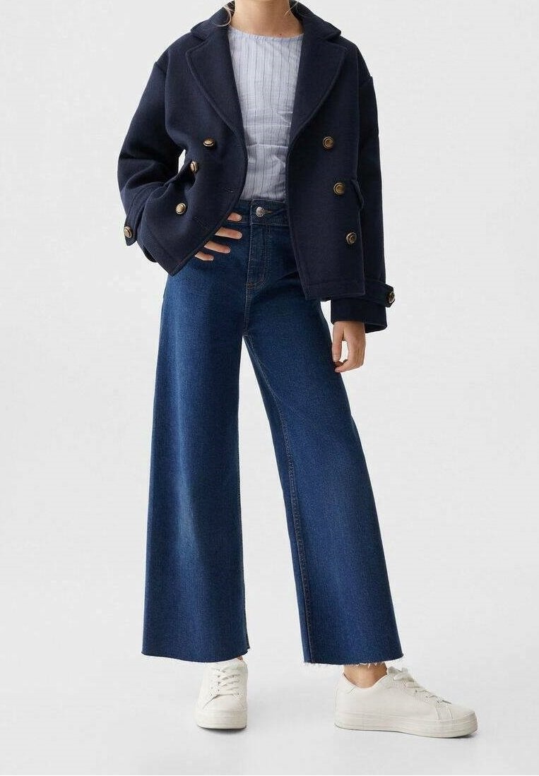 Marinblå peacoat med guldknappar, kombinerad med vida blå jeans och vita sneakers. Skjortan under har ljusa vertikala ränder.