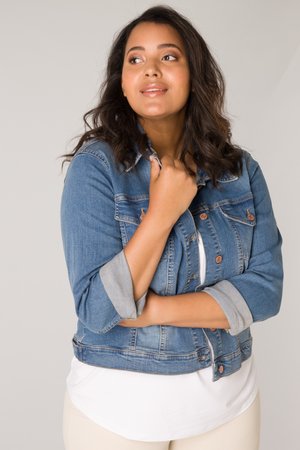 Jonge vrouw met donker haar die een denimjack met opgerolde mouwen draagt over een witte top, kijkt iets naar links met een zachte glimlach.