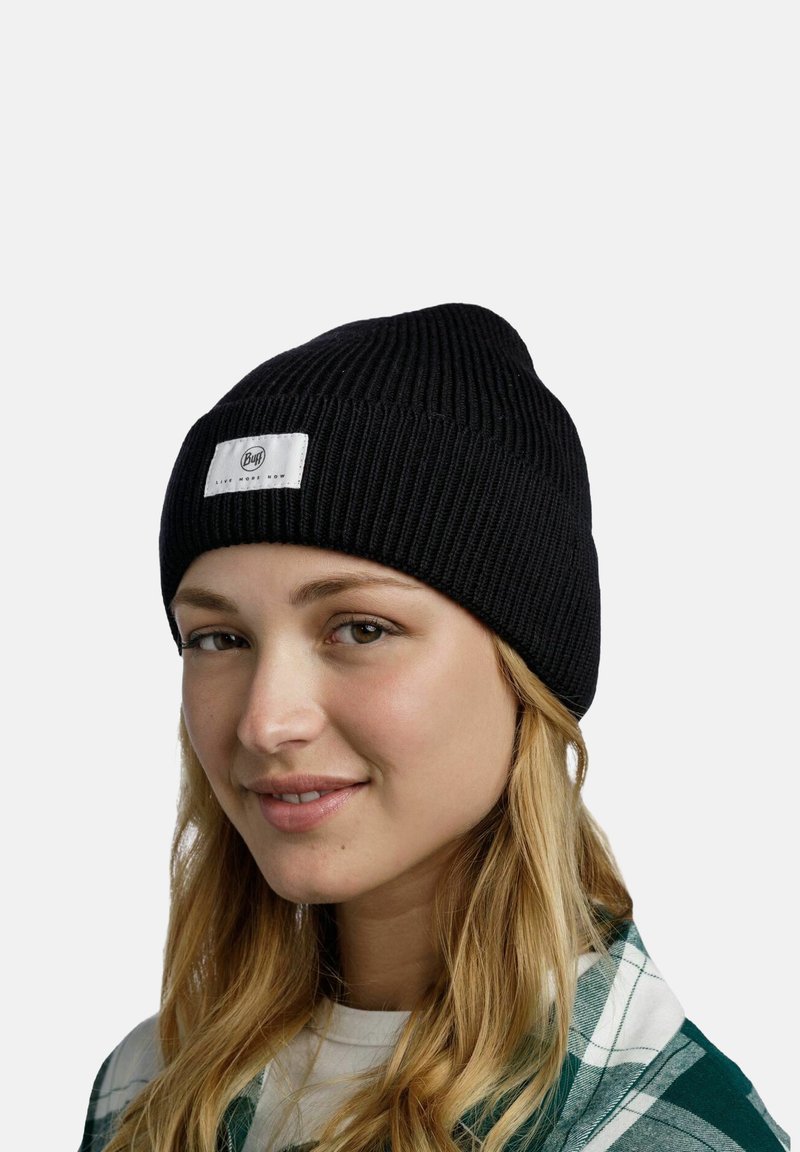 Gorro de punto acanalado negro con una etiqueta tejida blanca que presenta un logo circular. La tela parece suave y tiene un diseño elástico.