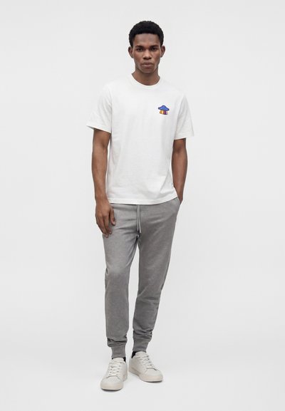 Άνδρας που φοράει άσπρο t-shirt με στρογγυλή λαιμόκοψη και μικρό πολύχρωμο γραφικό UFO, γκρι παντελόνι φόρμας με κορδόνι και άσπρα αθλητικά παπούτσια, στέκεται μπροστά σε απλό φόντο.