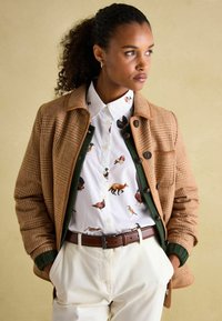 Brown houndstooth cardigan with green accents, white shirt featuring animal prints, beige trousers, and a brown belt. Textured material.