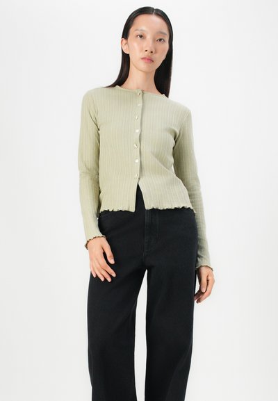 Ribbed long-sleeve cardigan in light green, featuring a rounded neckline, button closure, and scalloped hem. Paired with black wide-leg jeans.