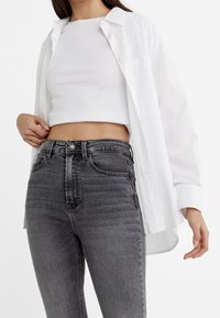 White cropped top layered under an open white button-up shirt; paired with high-waisted, dark gray denim jeans featuring a slight texture.