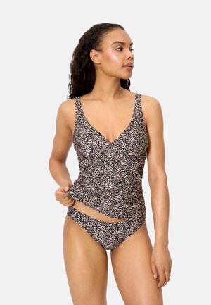 Mujer con un top de tankini de estampado a juego y braguitas de bikini, sujetando el borde del top y mirando de lado sobre un fondo liso.