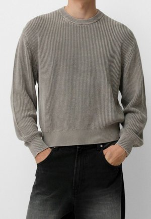 Man wearing a gray ribbed knit sweater with long sleeves and black jeans, hands tucked into front pockets.