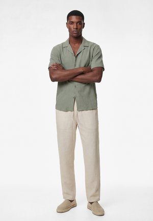 Mężczyzna stojący ze skrzyżowanymi ramionami, ubrany w jasnowierzchnią zieloną koszulę na guziki z krótkim rękawem, beżowe spodnie i beżowe buty wsuwane na białym tle.