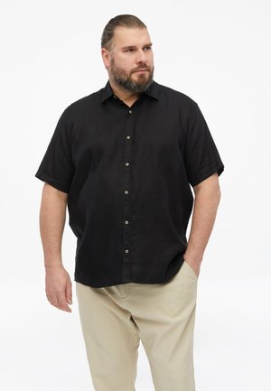 Mann mit Bart und zurückgebundenem Haar, trägt ein schwarzes kurzärmliges Hemd mit Knöpfen und beige Hose, eine Hand in der Tasche.