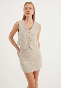 Beige tweed tvådelat set med en ärmlös väst som har tre knappar och en figurnära minikjol. Strukturerad tyg med ett rutmönster.