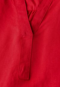 Tessuto di lino rosso con un design a V e una piccola piega. La texture è liscia, con cuciture visibili lungo i bordi e il colletto.