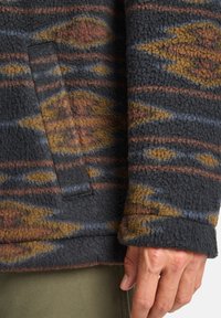 Gros plan sur une main près du poignet d'une veste en polaire noire avec des motifs géométriques marron, orange et bleu, et une poche latérale.