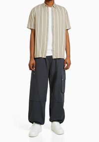 Vaaleanbeige lyhythihainen paita, jossa on valkoisia pystysuoria raitoja, yhdistettynä tummanharmaisiin cargo-housuihin, joissa on vetoketjulliset taskut ja väljä istuvuus. Valkoiset lenkkarit näkyvät.