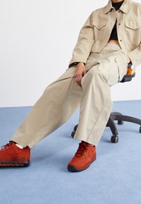 Osoba siedząca na biurowym krześle, ubrana w beżową kurtkę i spodnie, z jasnopomarańczowymi sneakersami na niebieskim dywanie.