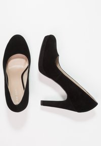 Černé semišové lodičky na vysokém podpatku s kulatou špičkou, výřezovými stranami a zužujícím se podpatkem. Interiér má hladké béžové podšívání.